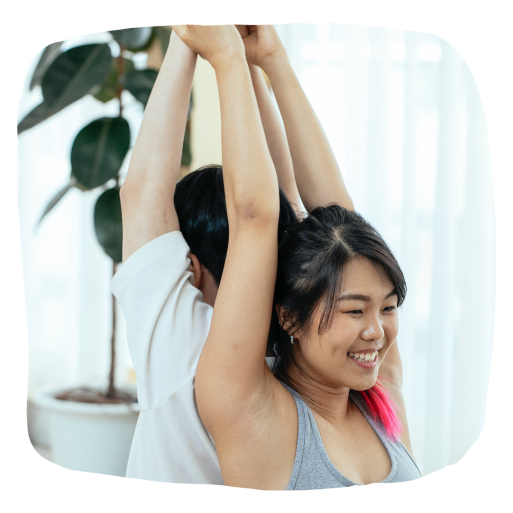 Two people (one of them smiling) positioned in a partner yoga pose with their hands stretched above their heads