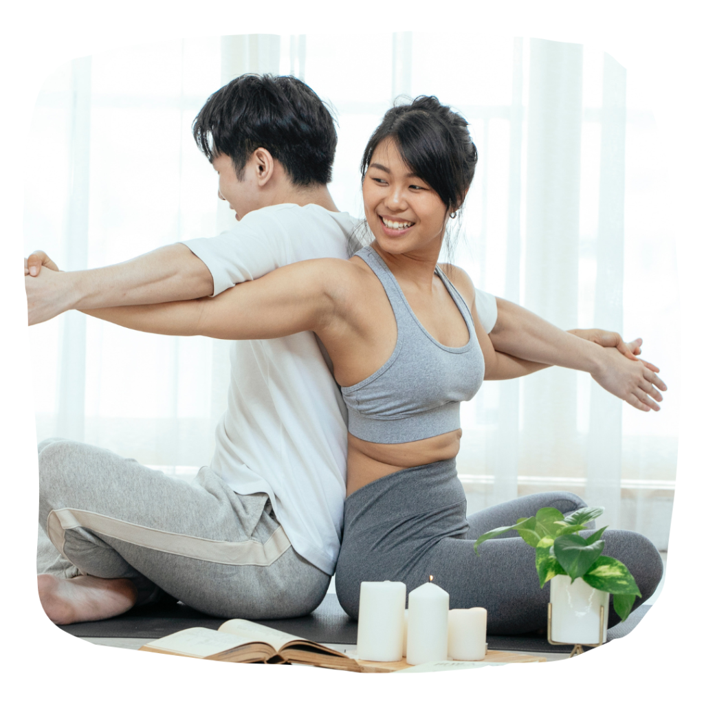 Two people sitting back to back (one of them smiling) positioned in a partner yoga pose with their arms stretched outward at their sides